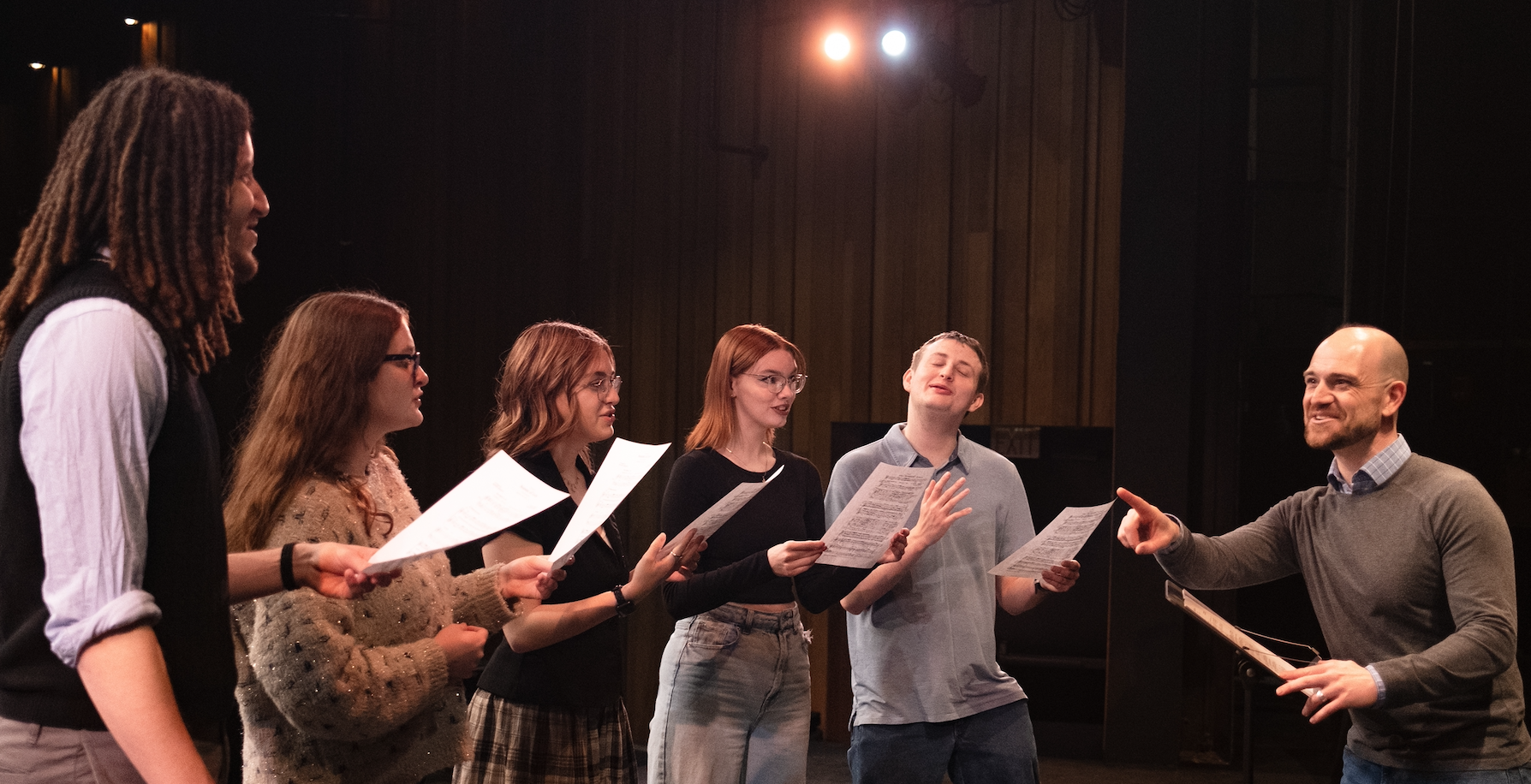 A group of 5 students singing while instructed by a professor.